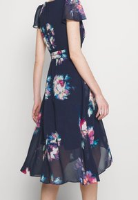 Navy kukkakuosiset mekko, jossa on läpinäkyvät lyhyet hihat ja vyötärönauha. Sisältää hulmuavan hameen, jossa on värikkäitä kukkakuviota ja kevyttä kangasta.