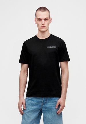 Ung mand med kort hår bærer en sort t-shirt med lille hvidt logo på brystet og lyseblå jeans, står foran en ensfarvet baggrund.