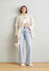 Béžový oversize blazer přes bílý cropped top, kombinovaný s světle modrými širokými džínami a bílými botami. Zahrnuje malou bílou kabelku.