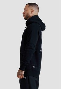 Uomo con taglio di capelli laterale e tatuaggio sul viso che indossa una felpa con cappuccio nera e pantaloni neri con piccole grafiche di lame bianche e rasoi, in piedi di lato.