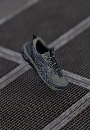 Szaro-czarny but sportowy ASICS GEL-Quantum 360 unoszący się nad metalową siatkową powierzchnią z teksturowaną podeszwą i wiązaniem na sznurowadła.