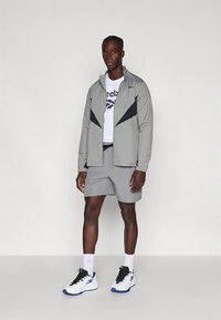 Άνδρας που φοράει γκρι jacket με μαύρες λεπτομέρειες, γκρι σορτς, άσσο T-shirt της Reebok, άσπρες κάλτσες και άσπρα sneakers με μαύρα κορδόνια και μπλε σόλες.