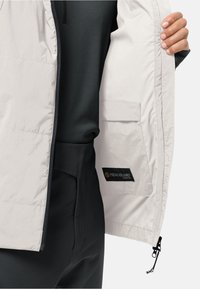 Personne tenant une veste beige ouverte montrant une poche intérieure étiquetée "MICROGUARD STRETCH" sur des vêtements noirs.