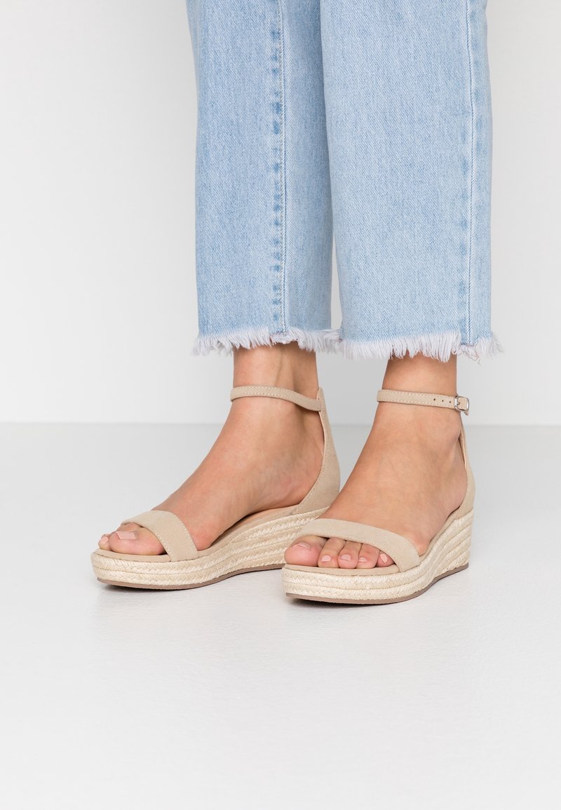 Sandales compensées beiges avec détail en corde style espadrille, bout ouvert, bride à la cheville et matériau en daim texturé. Assorties à un jean bleu clair à bords effilochés.