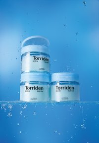 Tre transparente krukker med Torriden Multi Pad står i vand, med lyseblå etiketter og hvide låg, mens dråber sprøjter omkring dem.