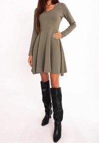 Femme portant une robe à manches longues vert olive avec une jupe évasée et des bottes noires hautes à talons sur fond blanc.