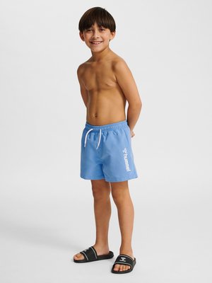 Uśmiechnięty chłopiec stoi boso, ubrany w jasnoniebieskie kąpielówki z białym sznurkiem i czarne sandały wsuwane na prostym białym tle.