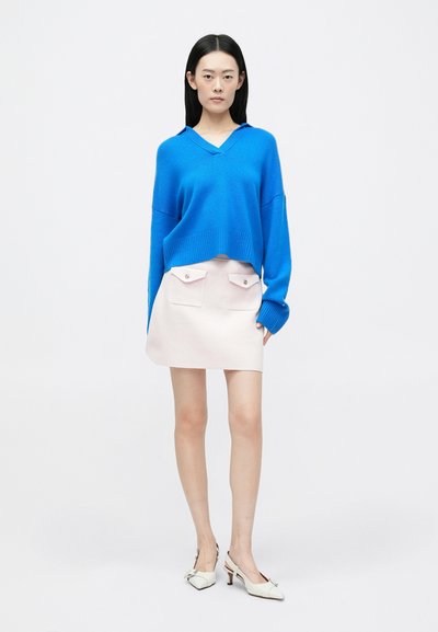 Niebieski, oversize'owy sweter dziany z dekoltem w serek, w połączeniu z różową mini spódnicą z dwoma przednimi kieszeniami. Białe sandały na słupku z paskiem za piętą.