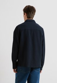 Person, der mit dem Rücken zur Kamera steht, trägt eine strukturierte dunkelblaue Jacke und Jeans auf einem hellen, einfarbigen Hintergrund.