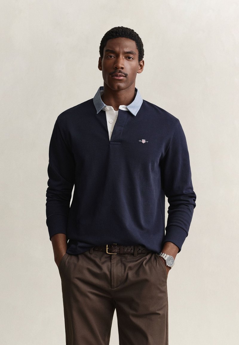 Uomo che indossa una polo blu navy a maniche lunghe con colletto azzurro, pantaloni marroni, cintura intrecciata scura e orologio da polso argentato, in piedi con le mani in tasca.