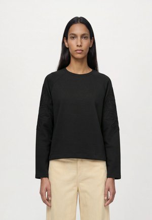 Femme aux longs cheveux foncés portant un haut noir à manches longues avec une broderie florale discrète sur les manches et un pantalon en velours côtelé beige, debout de manière neutre.