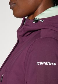 Veste imperméable violet avec capuche, dotée d'une doublure vert menthe, d'une poche zippée et d'un logo imprimé blanc sur l'épaule.