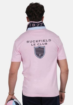 Homme portant un polo rose avec l'inscription "Ruckfield Le Club" et un logo de blason dans le dos, tenant un ballon de rugby, col relevé affichant le texte "1977".