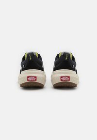 Sorte og gule sneakers med ruskinn og mesh-materialer, en kraftig hvit mellomsåle, strukturert brun yttersåle og rød Vans-logo på hælen.