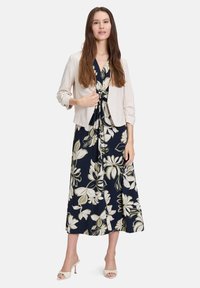Kwiatowa granatowa sukienka z białymi i zielonym wzorami, zwężona w talii, oraz jasnobeżowa krótka kurtka. Modelka nosi białe sandały na obcasie.
