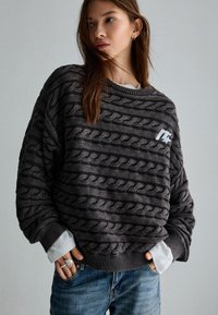 Overmaatse grijze trui met een gestructureerd kabelbreipatroon, geribde manchetten en een witte logo-accent. Draag het over een wit shirt, gecombineerd met een spijkerbroek.