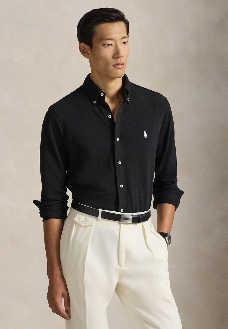 Μαύρο πουκάμισο με μακριά μανίκια και κουμπιά, με λευκό λογότυπο, συνδυασμένο με κρεμ πλισέ παντελόνια και μαύρη ζώνη με ασημί αγκράφα.