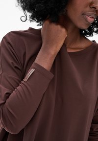 Bruine langemouwen top van zacht materiaal, met een label op de linkermouw, een ronde halslijn en een ontspannen pasvorm. Details in de textuur zijn zichtbaar.