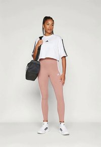 Bijela skraćena atletska majica s crnim prugama, svijetloružne tajice s bijelim bočnim prugama i crna teretna torba. Bijeli tenisice upotpunjuju odjevnu kombinaciju.