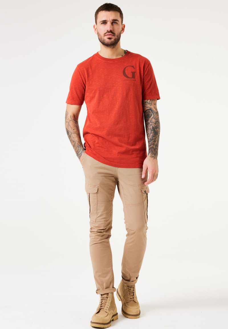 Camiseta roja de manga corta con cuello redondo y logo "G" negro. Combinada con pantalones cargo beiges con bolsillos laterales y dobladillos enrollados. Botas de cuerda marrones.