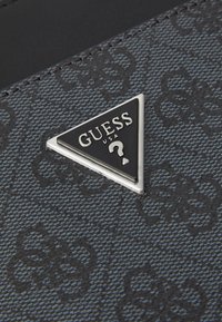 Tissu noir texturé présentant un motif répétitif. Logo triangulaire métallique avec "GUESS USA" et un détail en forme de point d'interrogation. Fini lisse.