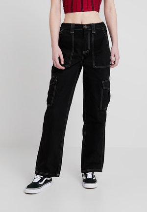 Personne portant un pantalon cargo noir avec des coutures blanches, des baskets noires et blanches, et un débardeur rouge court, debout sur un sol gris.