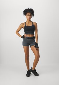 Černý sportovní crop top s tenkými ramínky, doplněný šedými vzorovanými šortkami. Obuté černé treninkové boty a rukavice na zápěstí.