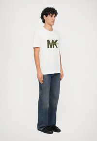 Vīrietis stāv, ģērbies baltā Michael Kors logotipa T-kreklā, brīvos zilos džinsos un melnās sporta kurpes fona balta krāsa.