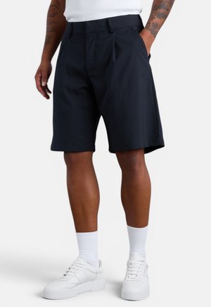 Mann trägt schwarze knielange Shorts, weiße Socken und weiße Sneakers, steht mit einer Hand in der Tasche vor einem einfarbigen Hintergrund.