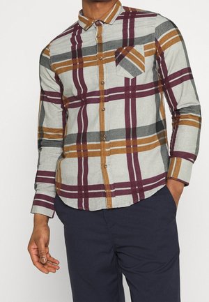 Man draagt een lang mouwen overhemd met een groot beige, bordeaux en grijs ruitpatroon, gecombineerd met donkerblauwe broek.