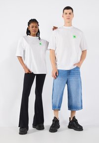 White oversized t-shirts feature green star graphics. The female model wears black flared pants; the male model wears blue knee-length denim shorts and black sneakers.