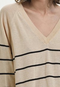 Pull beige à col en V avec des rayures horizontales noires, doté d'une texture douce et d'une coupe décontractée. Épaules tombantes remarquables et col côtelé.