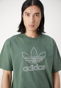 Oliwkowa bawełniana koszulka z krótkim rękawem, z dużym białym logo Adidas z trzema liśćmi i poziomymi paskami. Okrągły dekolt.