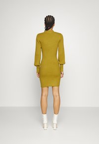Robe en maille côtelée jaune moutarde avec des manches longues et une silhouette ajustée, associée à des bottines à talons bloc blanches.