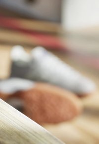 Sportovní boty v bílé a šedé barvě s texturovanou podrážkou, částečně rozostřené, na světle dřevěném povrchu, s rozmazaným hnědým objektem v blízkosti.