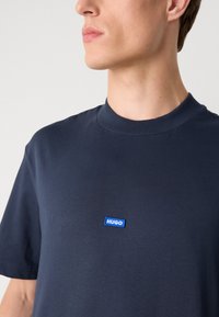 Sötétkék pamut póló kerek nyakkivágással és rövid ujjakkal, elöl kék téglalap alakú logópattal, amelyen a "HUGO" felirat látható.