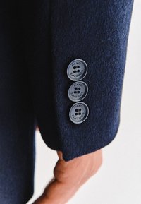 Mankiet marynarki w kolorze granatowym z trzema okrągłymi, szarymi guzikami, z których każdy jest oznaczony napisem "PAKO LORENTE". Materiał ma miękki, teksturowany wygląd.