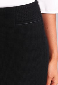 Nahaufnahme eines schwarzen strukturierten Rocks mit kleiner Schlitztasche vorne und einem Teil des Arms in einem weißen kurzärmeligen Oberteil.