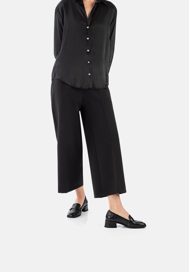Femme portant une chemise noire à manches longues boutonnée, un pantalon noir ample court et des mocassins noirs à talons, debout sur fond blanc.