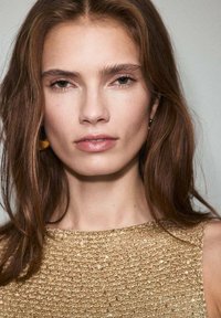 Top dorado texturizado con cuello redondo. La modelo tiene el cabello largo y ondulado de color castaño y lleva pendientes discretos. La piel se ve suave y natural.