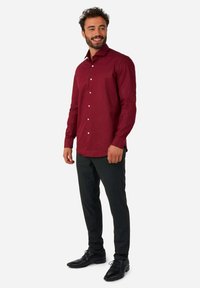 Camisa de manga larga burdeos con cuello clásico, parte delantera de botones y botones blancos, combinada con pantalones oscuros y zapatos negros.