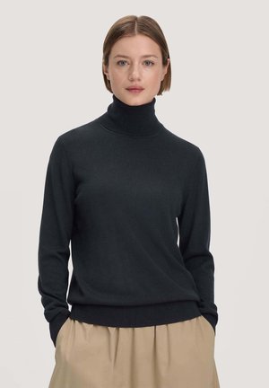 Jonge vrouw met kort haar, gekleed in een zwarte coltrui en beige broek, staand voor een effen lichte achtergrond.