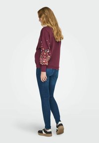 Felpa bordeaux con ricami floreali sulle maniche, polsini a coste e colletto tondo, abbinata a jeans skinny blu e sneakers bianco e nero.