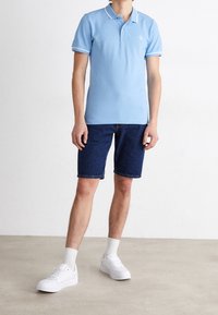 Jasnoniebieska koszulka polo z białym kołnierzykiem i akcentami na rękawach, w połączeniu z ciemnoniebieskimi szortami dżinsowymi. Białe sneakersy i jasne skarpetki dopełniają stylizację.