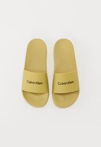 Sandale galbene din material neted, cu o curea lată pe care este inscripționat "Calvin Klein" cu litere negre. Talpă plată, design simplu.