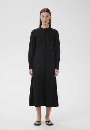 Kobieta stojąca przodem, ubrana w czarną midi sukienkę na długi rękaw z guzikami oraz czarne sandały, na białym tle.