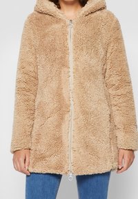 Beige, fluffy zip-up hættejakke båret over blå jeans, vist fra skuldre til øvre lår.