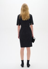 Černé úzké šaty s krátkými rukávy, délka ke kolenům a zip na zadní straně. Nosené s černými ponožkami a botami, drží malou černou kabelku.