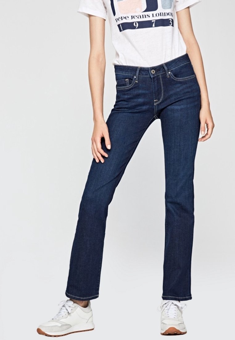 Personne portant un jean droit bleu foncé, un t-shirt blanc avec un motif graphique et des baskets blanches, debout avec une main posée sur la cuisse.