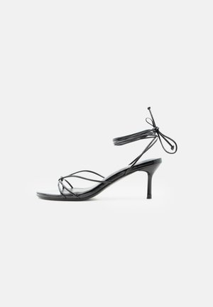 Fekete, pántos, magas sarkú szandál vékony bokapántokkal és közepes magasságú tűsarkúval, fehér háttéren.